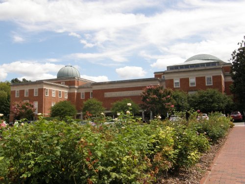 A brick building with a dome, surrounded by blooming flowers and trees under a partly cloudy sky.
