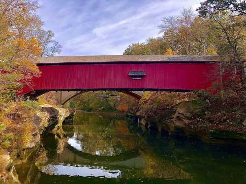 A red covered bridge spans a calm river, surrounded by autumn foliage and a clear blue sky.