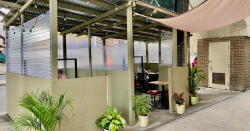 Outdoor dining area with clear plastic barriers, plants, and a shaded entrance, located on a city street.