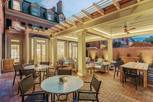 Outdoor dining area with tables and chairs under a pergola, surrounded by brick walls and greenery at dusk.