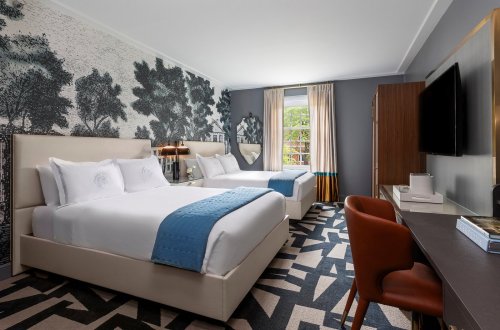 A modern hotel room featuring two beds, a desk, and stylish decor with a nature-themed wallpaper.