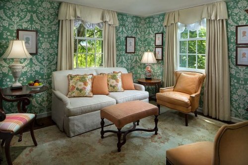 Cozy living room with floral wallpaper, a white sofa, orange armchair, and natural light from windows.