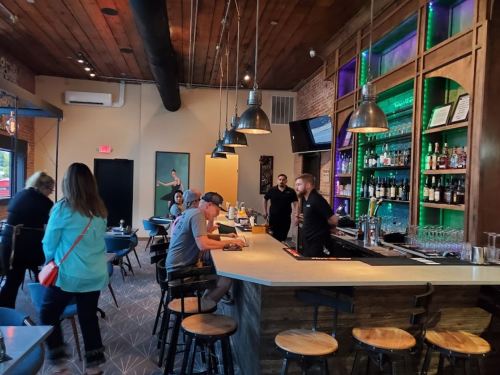 A lively bar scene with patrons at the counter, staff serving drinks, and colorful lighting in a modern setting.