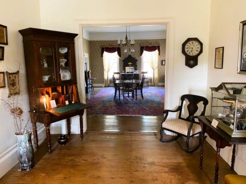 A hallway leading to a dining room, featuring vintage furniture, a patterned wallpaper, and a colorful area rug.