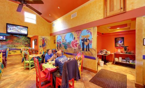 Colorful restaurant interior with vibrant murals, wooden chairs, and patrons dining at tables. A TV is mounted on the wall.