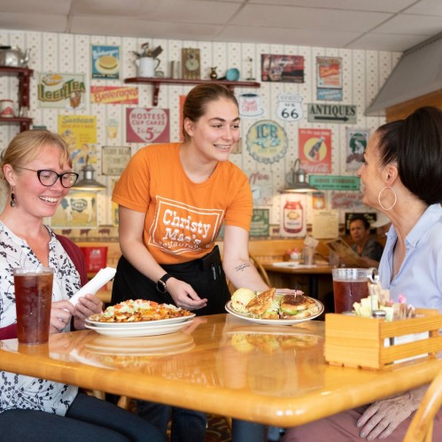 Three women enjoy a meal at a cozy restaurant, with vintage decor and plates of food on the table.