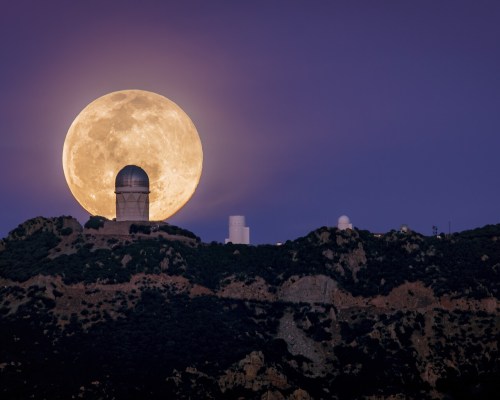 A large full moon rises behind a mountain observatory, casting a serene glow over the landscape.