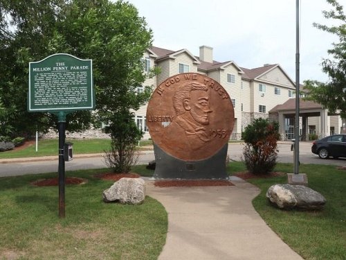 A large penny monument with a plaque nearby, set in a grassy area in front of a building.