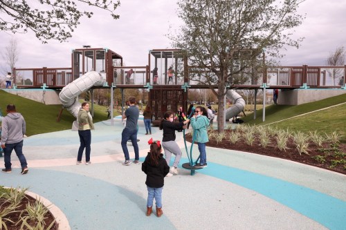 Children play on a colorful playground with slides and climbing structures, surrounded by greenery and adults nearby.