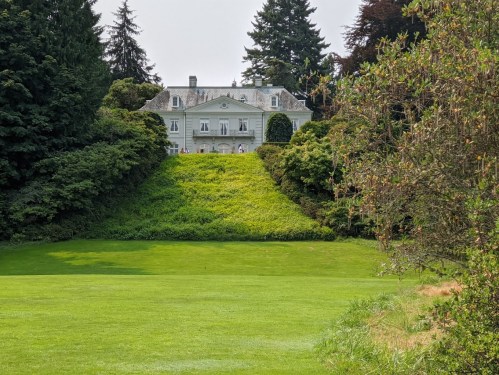 A large, elegant mansion surrounded by lush greenery and trees, set on a hillside with a manicured lawn in the foreground.