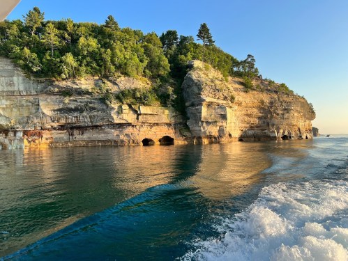 A scenic view of a rocky cliff covered in greenery, reflecting in calm waters under a clear blue sky.