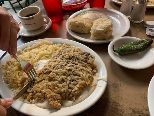 A plate of fried fish with gravy, hash browns, biscuits, and a jalapeño, with a cup of coffee in the background.