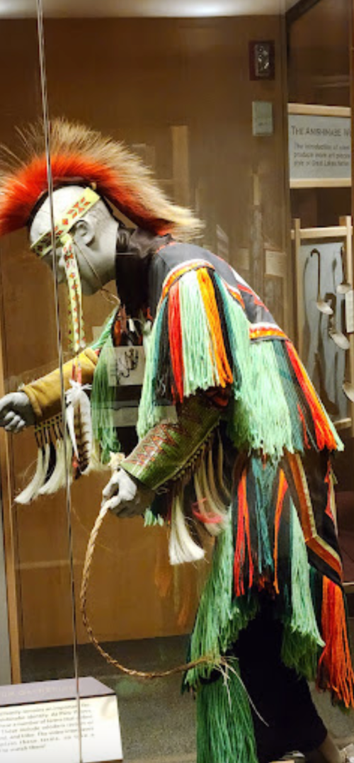 A colorful traditional costume displayed in a museum, featuring vibrant fringe and a feathered headdress.