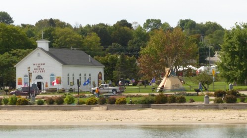 A museum building with flags, surrounded by trees and a teepee, near a sandy beach and water.