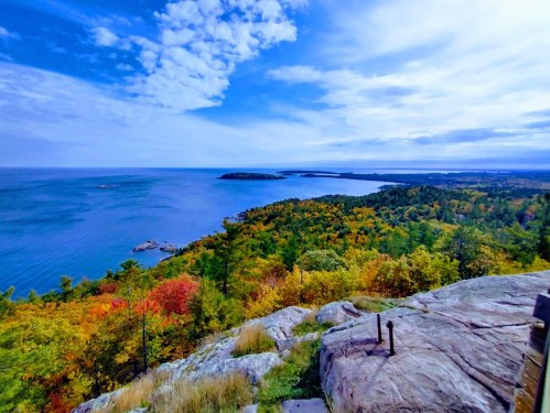 A scenic view of a colorful autumn landscape overlooking a calm blue lake and distant islands under a partly cloudy sky.