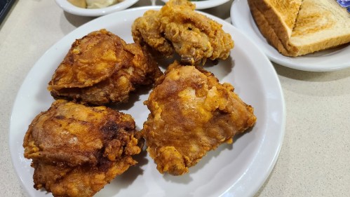 A plate of crispy fried chicken pieces with toast and side dishes in the background.