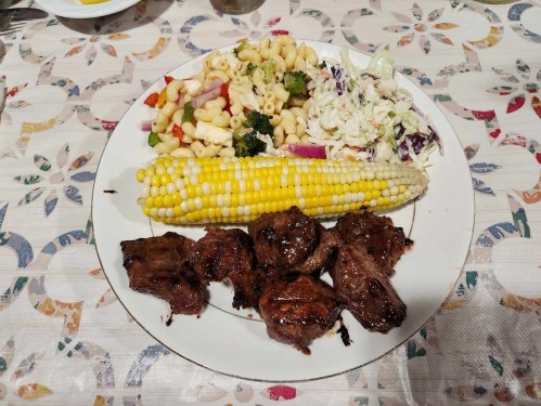 A plate with grilled steak, corn on the cob, pasta salad with vegetables, and coleslaw on a patterned tablecloth.