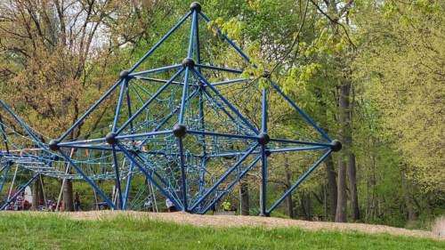 A blue geometric climbing structure in a park, surrounded by trees and grass.