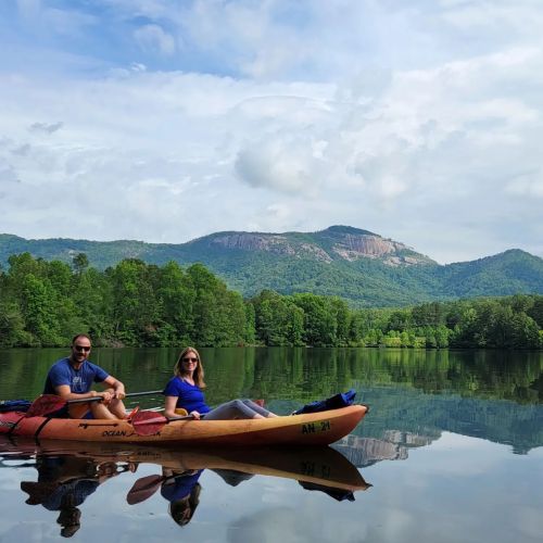 Two people sit in a kayak on a calm lake, surrounded by lush greenery and mountains under a partly cloudy sky.