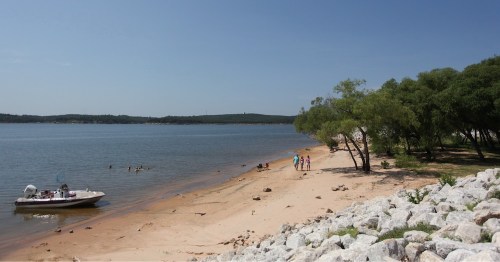 A sandy beach by a calm lake, with people walking and swimming, and a boat anchored nearby under a clear blue sky.