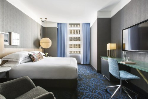 Modern hotel room featuring a king bed, desk, chair, and large window with blue curtains. Stylish decor and lighting.