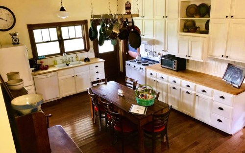 A cozy kitchen with a wooden dining table, white cabinets, pots hanging, and a window letting in natural light.