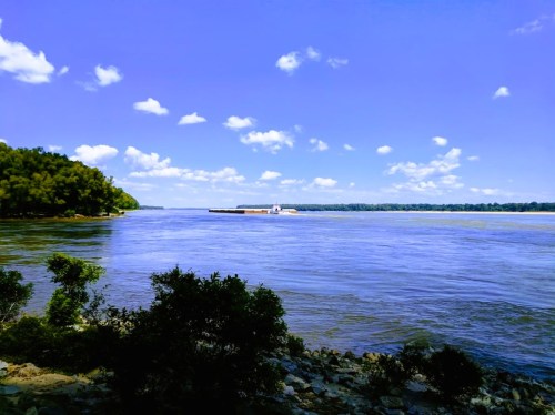 A serene river scene with a barge on the water, surrounded by lush greenery and a clear blue sky with fluffy clouds.