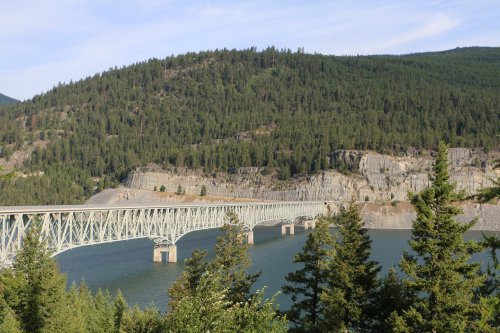 A metal bridge spans a river, surrounded by lush green hills and rocky cliffs under a clear blue sky.