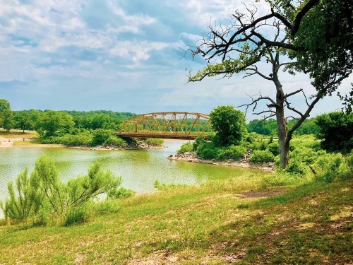 A scenic view of a bridge over a calm river, surrounded by lush greenery and a cloudy sky.