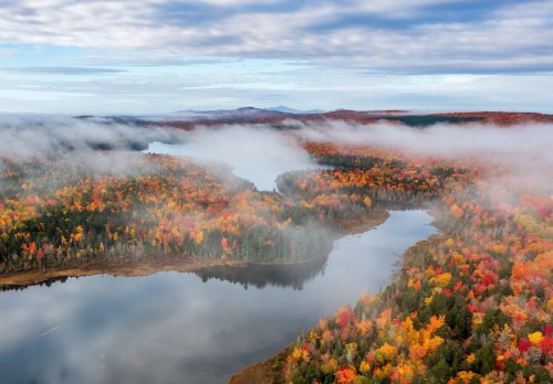 Aerial view of a colorful autumn landscape with a river, surrounded by mist and vibrant fall foliage.