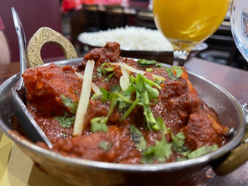 A bowl of spicy chicken curry garnished with cilantro and ginger, served with rice and a drink in the background.