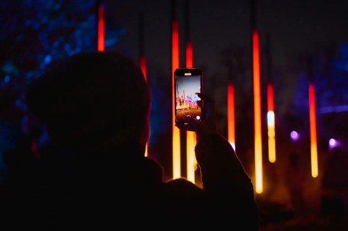 A person holds a phone, capturing a colorful light display with tall, glowing pillars at night.