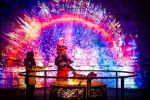 A family enjoys a colorful light display while gathered around a fire pit, with one child on an adult's shoulders.