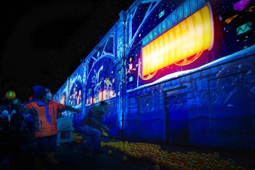Children play near a brightly lit mural, with colorful projections and a vibrant atmosphere in a dark setting.