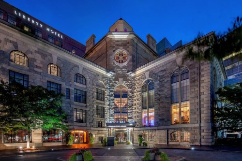 Historic building at dusk, featuring large windows and a prominent clock, surrounded by greenery and modern architecture.