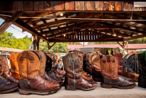 A display of various styles of leather cowboy boots on a wooden table under a rustic shelter.