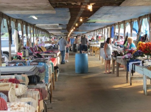 A bustling market scene with people browsing tables filled with various goods under a covered area.