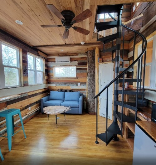Cozy interior of a tiny house featuring a spiral staircase, wooden walls, a blue sofa, and a small coffee table.