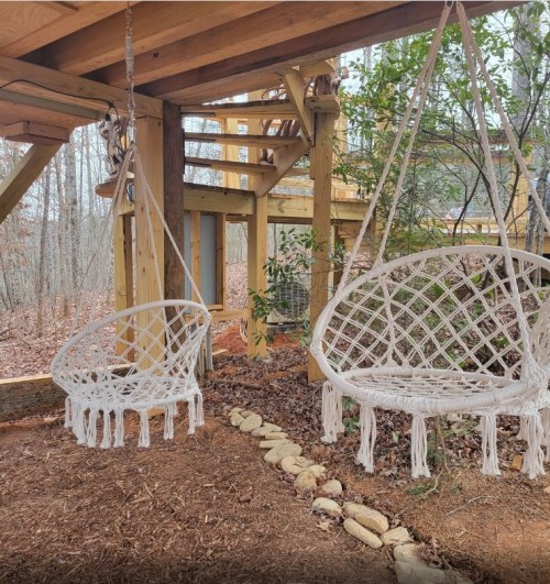 Two hanging macramé chairs under a wooden structure, surrounded by trees and gravel pathways.