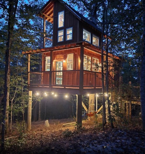 A modern treehouse elevated among trees, illuminated at dusk with large windows and a wooden deck.
