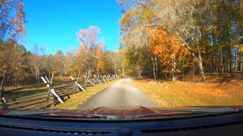 A scenic drive along a tree-lined road with vibrant autumn foliage and a wooden fence on the left.