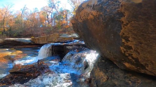 A close-up view of a rocky stream with cascading water, surrounded by autumn foliage and trees.