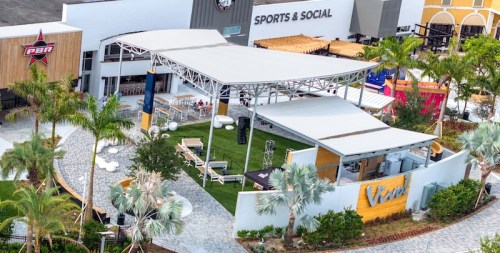 Aerial view of a vibrant outdoor venue with a stage, seating, and palm trees, surrounded by modern buildings.