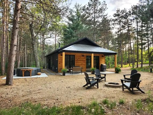 A modern cabin surrounded by trees, featuring a patio, fire pit, and outdoor seating area.