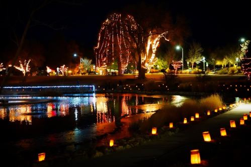 A serene night scene with a pond reflecting colorful lights and lanterns along a path, surrounded by illuminated trees.