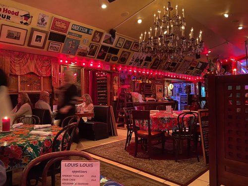 Cozy restaurant interior with red lighting, floral tablecloths, and vintage decor, featuring chandeliers and framed art.