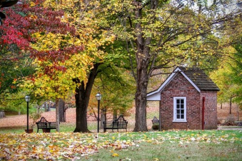 A small brick building surrounded by colorful autumn trees and benches, with fallen leaves covering the ground.