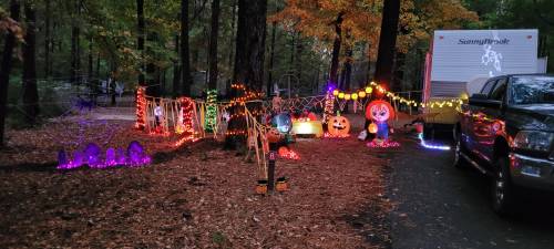 A festive outdoor scene with colorful Halloween decorations, including lights, pumpkins, and spooky figures among autumn trees.