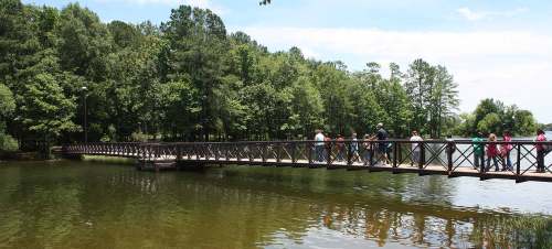 A wooden bridge spans a calm lake, with people walking and lush green trees lining the shore under a clear blue sky.