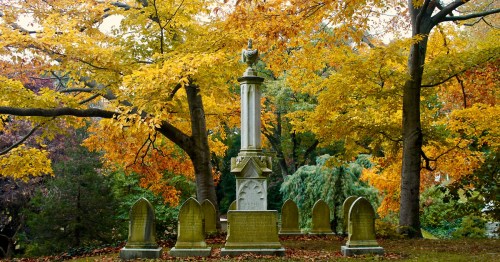 A historic monument surrounded by colorful autumn trees and several gravestones in a serene cemetery setting.